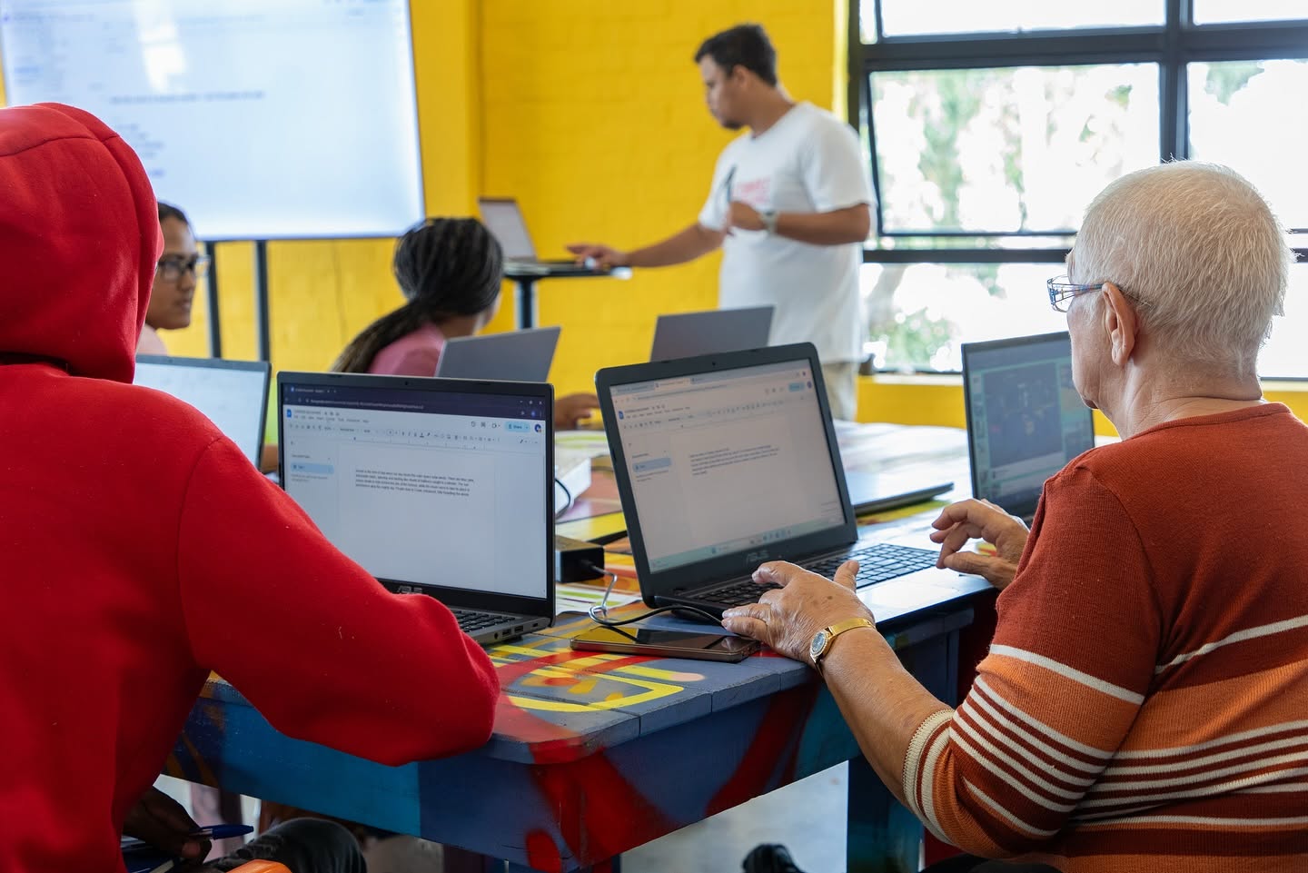 Adults working on laptops in a bright classroom while a facilitator presents